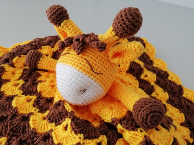 May include: A crocheted giraffe soft toy with brown spots and a yellow and brown blanket. The giraffe is lying on the blanket with its head resting on the blanket.
