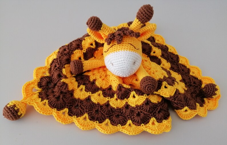 May include: A crocheted giraffe security blanket with brown and yellow stripes. The giraffe has a white face and brown spots. The blanket has a scalloped edge.