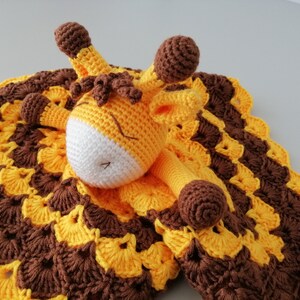 May include: A crocheted giraffe security blanket with a brown and yellow pattern. The giraffe is lying on the blanket with its head resting on the blanket.