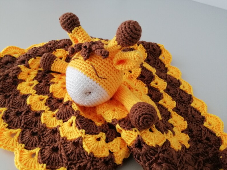 May include: A crocheted giraffe security blanket with a brown and yellow pattern. The giraffe is lying on the blanket with its head resting on the blanket.