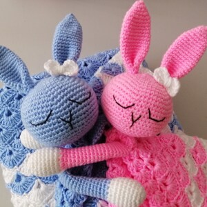 May include: Two crocheted bunny plushies, one blue and one pink, with white bows, are lying on a white and blue crocheted blanket.
