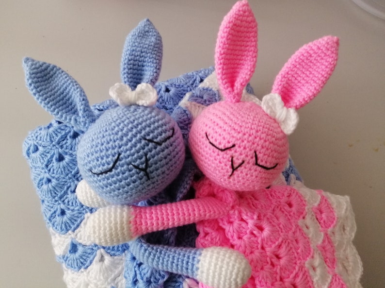 May include: Two crocheted bunny plushies, one blue and one pink, with white bows, are lying on a white and blue crocheted blanket.