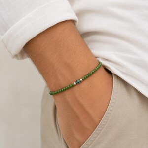 Op de afbeelding: Een kralenarmband met kleine, ronde, donkergroene kralen in een doorlopende lijn. De armband is geaccentueerd met een enkele, gefacetteerde, donkergroene kristallen kraal, geflankeerd door twee kleine, gepolijste zilverkleurige metalen tussenkralen.
