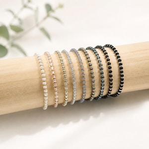 May include: A collection of nine thin beaded bracelets arranged in a row. Each bracelet features small, uniform round beads in a gradient of colors ranging from white and cream to grey, blue, and black, interspersed with tiny metallic gold spacers.