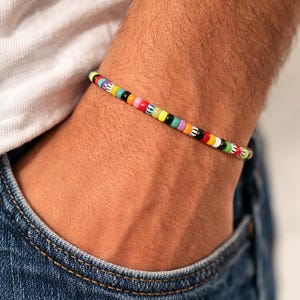 Könnte beinhalten: Ein farbenfrohes Perlenarmband mit einer Vielzahl kleiner, runder Perlen in Regenbogenfarben. Das Armband wird am Handgelenk getragen, mit einem weißen Hemd und einer blauen Jeans im Hintergrund. Die Perlen haben einen Durchmesser von ca. 0,6 cm.