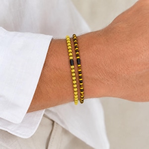 Könnte beinhalten: Zwei Perlenarmbänder, die zusammen getragen werden. Ein Armband besteht aus kleinen, einheitlichen gelben Perlen mit einer schwarzen zylindrischen Perle. Das zweite Armband besteht aus braunen und gelben Tigeraugenperlen mit einer zentralen schwarzen zylindrischen Perle.