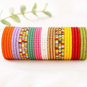 Op de afbeelding: Een verzameling gestapelde armbanden gemaakt van kleine, ronde, kleurrijke glaskralen. De set bevat effen banden in oranje, lavendel, groen, roze, wit, goud, geel, rood en olijfgroen, naast strengen met veelkleurige kralenpatronen.