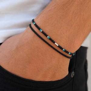 Peut inclure: Deux bracelets superposés composés de petites perles noires. L'un est entièrement noir, tandis que l'autre alterne des perles noires avec de petites perles turquoise et deux accents cylindriques de couleur argentée, formant un design perlé.
