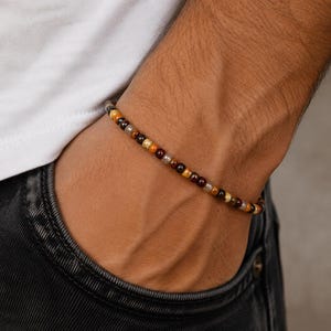 Könnte beinhalten: Ein Perlenarmband mit einer Mischung aus braunen, gelben und klaren Perlen. Das Armband wird am Handgelenk getragen. Die Perlen sind klein und rund und bilden ein schlichtes, elegantes Design.
