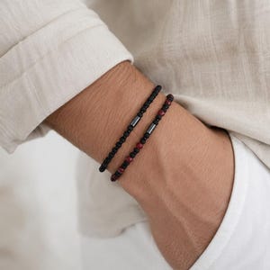 Op de afbeelding: Twee kralenarmbanden die samen worden gedragen. De ene armband heeft kleine zwarte kralen met een centrale metalen cilinder. De tweede armband bestaat uit afwisselende zwarte en donkerrode kralen, ook met een centrale metalen cilinderkraal.