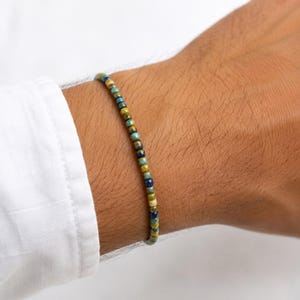 May include: A beaded bracelet featuring small, round beads in blue, green, and gold hues. The bracelet is displayed on a wrist, highlighting the colours and texture of the beads. The bracelet is a piece of jewellery.