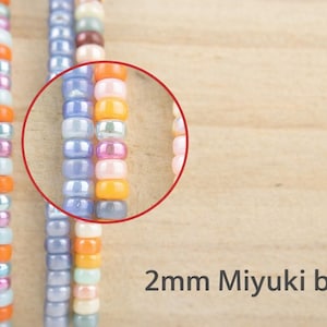 K&ouml;nnte beinhalten: Str&auml;nge aus 2mm Miyuki-Perlen in verschiedenen Farben wie Hellblau, Orange, Pink und Violett. Ein roter kreisf&ouml;rmiger Umriss hebt einen Abschnitt der Perlen hervor und zeigt ihre zylindrische Form und gl&auml;nzende Oberfl&auml;che. Der Text 2mm Miyuki beads ist sichtbar.