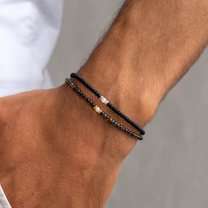 May include: Two beaded bracelets on a wrist. The top bracelet is black with a small white gem. The bottom bracelet is brown with gold and white beads. The bracelets are worn on a person's arm.