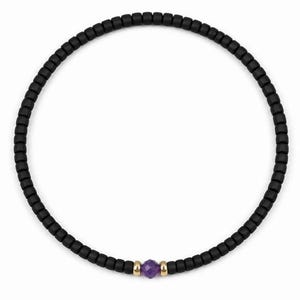 May include: Black beaded necklace featuring a central purple gemstone and gold-coloured accents. The necklace is made of small, square black beads. The gemstone is a deep purple colour. The necklace is shown against a white background.