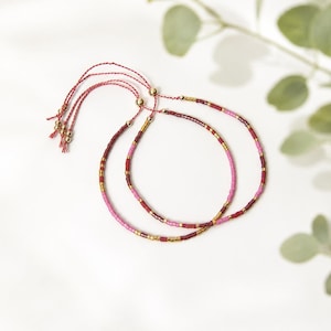 May include: Two adjustable bracelets crafted from thin red cord, featuring a repeating pattern of small cylindrical beads in shades of pink, deep red, and metallic gold. Each bracelet ends with a sliding knot mechanism and small gold-toned beads.