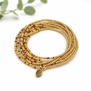 May include: A multi-strand wrap bracelet composed of small, round beads in alternating shades of metallic gold, tan, and dark brown. The bracelet features a circular, textured gold-toned metal charm with a spiral pattern hanging from the end.