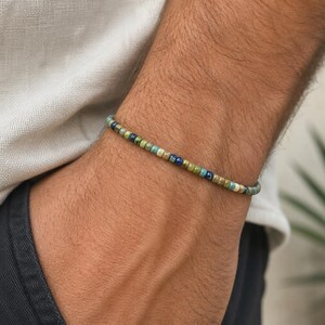 May include: A beaded bracelet with a mix of green, blue, and tan beads. The bracelet is worn on a wrist, with a neutral-colored shirt and dark pants in the background. The bracelet is a simple, minimalist design.