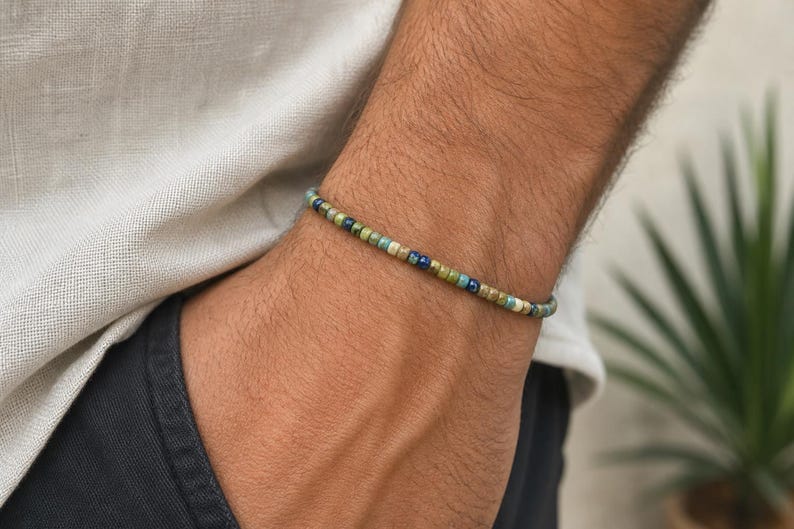 May include: A beaded bracelet with a mix of green, blue, and tan beads. The bracelet is worn on a wrist, with a neutral-colored shirt and dark pants in the background. The bracelet is a simple, minimalist design.