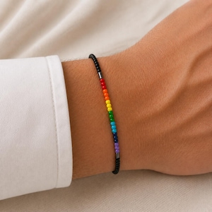 Op de afbeelding: Een dunne armband met een reeks kleine zwarte kralen afgewisseld met een levendig regenboogkleurig gedeelte. Het regenboogsegment bevat individuele rode, oranje, gele, groene, lichtblauwe, donkerblauwe en paarse kralen, gescheiden door kleine metalen tussenstukjes.