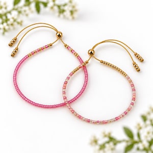 Könnte beinhalten: Zwei verstellbare Armbänder mit dünnen beigen Kordeln und kleinen Perlen. Ein Armband hat leuchtend rosa und goldene Perlen, das andere eine Mischung aus rosa, transparenten und goldenen Perlen. Beide haben verschiebbare goldene Metallperlen.