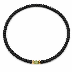 May include: Black beaded necklace featuring a central peridot gemstone and gold-coloured accents. The necklace is made of small, round, matte black beads. The peridot is a light green, faceted gemstone.