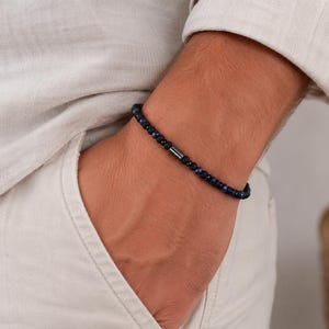 Könnte beinhalten: Ein Perlenarmband mit kleinen, runden, dunkelblauen und schwarzen Steinen in einem sich wiederholenden Muster. Das Armband enthält eine einzelne, glatte, zylindrische Metallperle in Dunkelgrau als zentrales Akzentstück.