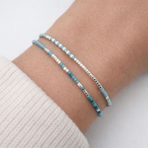 Op de afbeelding: Twee delicate armbanden met kralen in blauwe en zilveren tinten worden om een pols gedragen. De armbanden hebben kleine, gelijkmatig verdeelde kralen, wat zorgt voor een minimalistisch en elegant ontwerp. De pols is gedeeltelijk zichtbaar, met een witte mouw op de achtergrond.