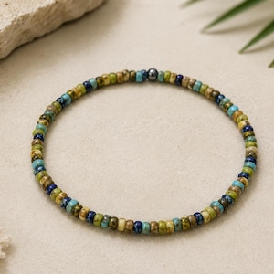 May include: A beaded bracelet featuring small, round beads in shades of blue, green, and tan. The bracelet has a dark gray metal accent bead. The jewelry is displayed on a neutral surface with a natural, earthy aesthetic.