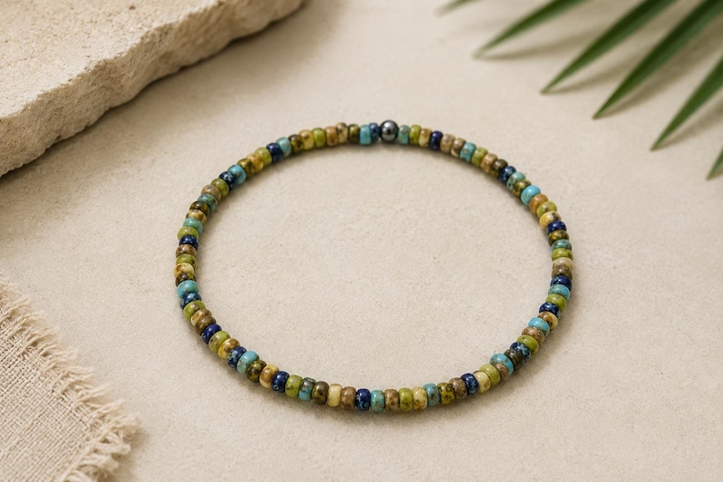 May include: A beaded bracelet featuring small, round beads in shades of blue, green, and tan. The bracelet has a dark gray metal accent bead. The jewelry is displayed on a neutral surface with a natural, earthy aesthetic.