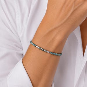 May include: A beaded bracelet with turquoise beads and silver-coloured cylindrical and round beads. The bracelet is worn on a wrist, against a white shirt.