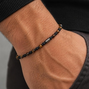 Peut inclure: Bracelet de perles noires, marron et dorées, orné d'une perle cylindrique argentée. Le bracelet est porté au poignet. On aperçoit une manche sombre et un pantalon gris. Les perles mesurent environ 0,5 cm de diamètre.