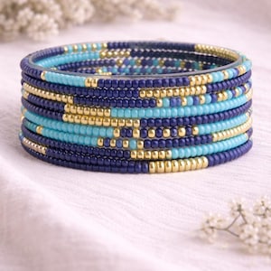 Könnte beinhalten: Ein Stapel Perlenarmbänder in Blau- und Goldtönen. Die Armbänder weisen eine Mischung aus dunkelblauen, hellblauen und goldenen Perlen auf, wodurch ein strukturiertes und farbenfrohes Accessoire entsteht. Die Armbänder sind aufeinander gestapelt und präsentieren das Design.