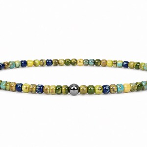 May include: A beaded bracelet featuring small, colorful beads in shades of green, blue, and yellow. A single, dark gray bead is centered in the design. The bracelet is strung on an elastic cord.