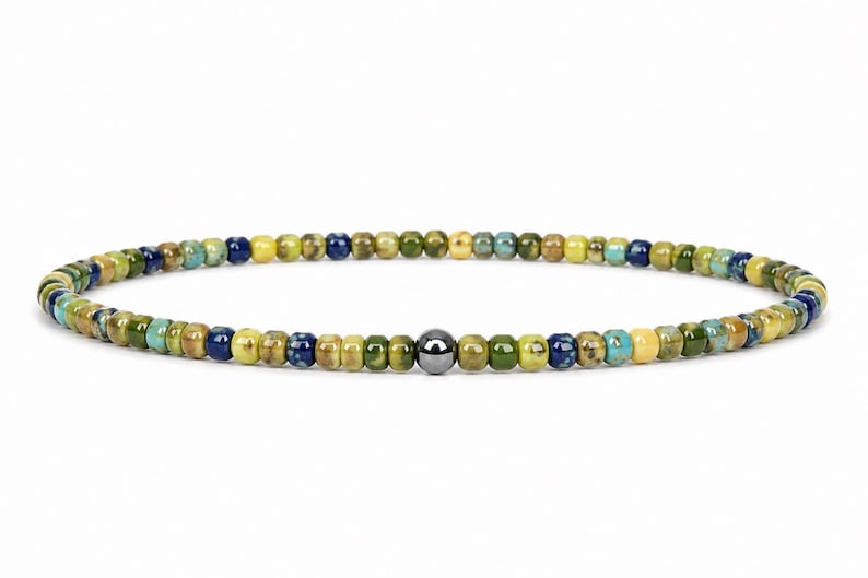May include: A beaded bracelet featuring small, colorful beads in shades of green, blue, and yellow. A single, dark gray bead is centered in the design. The bracelet is strung on an elastic cord.