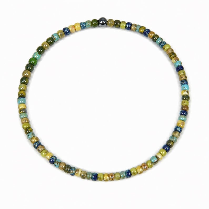 May include: A beaded bracelet featuring small, colorful beads in shades of green, blue, yellow, and brown. The beads are strung together to form a circular shape. A single, dark metallic bead is centered on the bracelet.