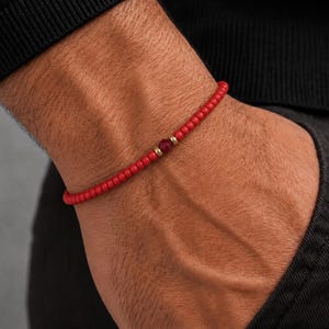 Könnte beinhalten: Rotes Perlenarmband mit einer zentralen dunkelroten Perle und goldfarbenen Akzenten. Das Armband wird am Handgelenk getragen, wobei der Arm und ein Teil eines dunkelfarbigen Hemdes sichtbar sind.