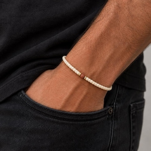 Könnte beinhalten: Cremefarbenes Perlenarmband mit einer rötlich-braunen Akzentperle und goldfarbenen Abstandshaltern. Das Armband wird am Handgelenk getragen, die Person trägt ein schwarzes T-Shirt und dunkle Jeans.