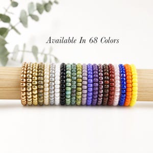 May include: A collection of eighteen elastic bracelets made of small, round, glossy beads in various colors including gold, silver, white, black, green, purple, brown, red, pink, blue, orange, and yellow, with the text Available In 68 Colors.