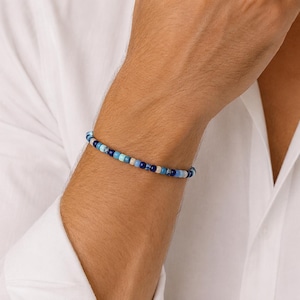Könnte beinhalten: Ein Armband mit kleinen, runden Perlen in Blau- und Weißtönen. Das Armband wird am Handgelenk einer Person getragen, die ein weißes Hemd trägt. Die Perlen sind in verschiedenen Blautönen gehalten und bilden ein schlichtes Accessoire.