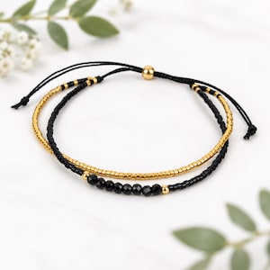 Op de afbeelding: Een dubbelstrengs armband bestaande uit een streng metallic gouden kraaltjes en een streng zwarte kraaltjes, met een centraal gedeelte van gefacetteerde zwarte kralen, kleine goudkleurige metalen kraaltjes en verstelbare zwarte koordjes.
