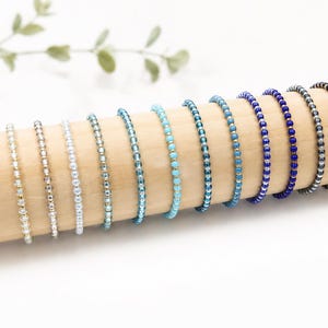 Könnte beinhalten: Eine Sammlung von elf dünnen Perlenarmbändern, die in einer Reihe angeordnet sind und einen Farbverlauf von Weiß und Gold bis hin zu Hellblau, Türkis, Königsblau, Grau und Schwarz aufweisen, jeweils aus kleinen, gleichmäßigen Perlen.