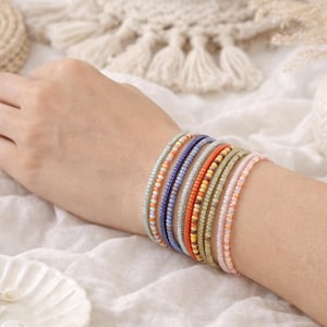 K&ouml;nnte beinhalten: Ein Stapel aus acht d&uuml;nnen Perlenarmb&auml;ndern am Handgelenk. Die Armb&auml;nder bestehen aus kleinen, gleichm&auml;&szlig;igen Glasperlen in verschiedenen Farben, darunter Hellblau, Orange, Dunkelblau, Silber, leuchtendes Orange, Gelb, Olivgr&uuml;n und zartes Rosa.