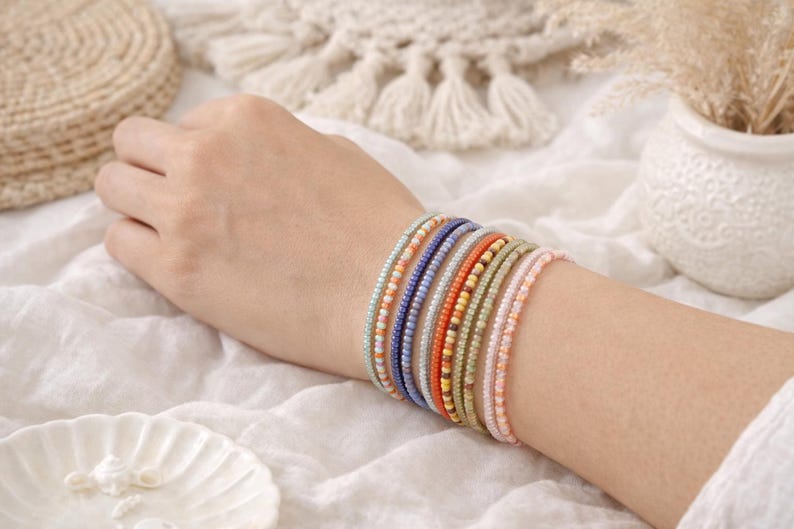 K&ouml;nnte beinhalten: Ein Stapel aus acht d&uuml;nnen Perlenarmb&auml;ndern am Handgelenk. Die Armb&auml;nder bestehen aus kleinen, gleichm&auml;&szlig;igen Glasperlen in verschiedenen Farben, darunter Hellblau, Orange, Dunkelblau, Silber, leuchtendes Orange, Gelb, Olivgr&uuml;n und zartes Rosa.