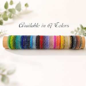Op de afbeelding: Een horizontale houten displaystang met een lange rij kleine, ronde, veelkleurige glazen kraaltjes gerangschikt in een kleurenspectrum. De tekst "Available in 67 Colors" staat in zwarte schrijfletters boven de kralen op de witte achtergrond.