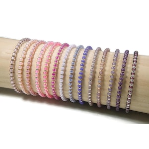 Könnte beinhalten: Eine Sammlung von Armbändern mit Perlen in verschiedenen Farben, darunter Rosa, Lila und Weiß, auf einem Holzstab präsentiert. Jedes Armband hat kleine, farbige Perlen und goldfarbene Akzente. Die Armbänder sind zart und scheinen handgefertigt zu sein.