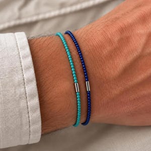 Peut inclure: Deux bracelets fins en perles portés ensemble. L'un est composé de petites perles turquoise et l'autre de petites perles bleu foncé. Chaque bracelet comporte une perle d'accent cylindrique en métal argenté, lisse et brillante.