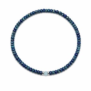 Op de afbeelding: Een kralenketting met kleine, ronde, donkerblauwe kralen met een gespikkeld patroon. Een lichtblauwe edelsteen is gecentreerd op de ketting, vastgehouden door zilverkleurig metaal. De ketting wordt op een witte achtergrond getoond.