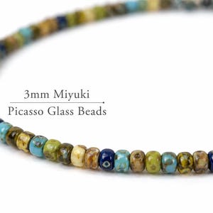 May include: A close-up of a strand of 3mm Miyuki Picasso glass beads. The beads are a mix of colors, including blue, green, yellow, and brown. The text "3mm Miyuki Picasso Glass Beads" is visible.