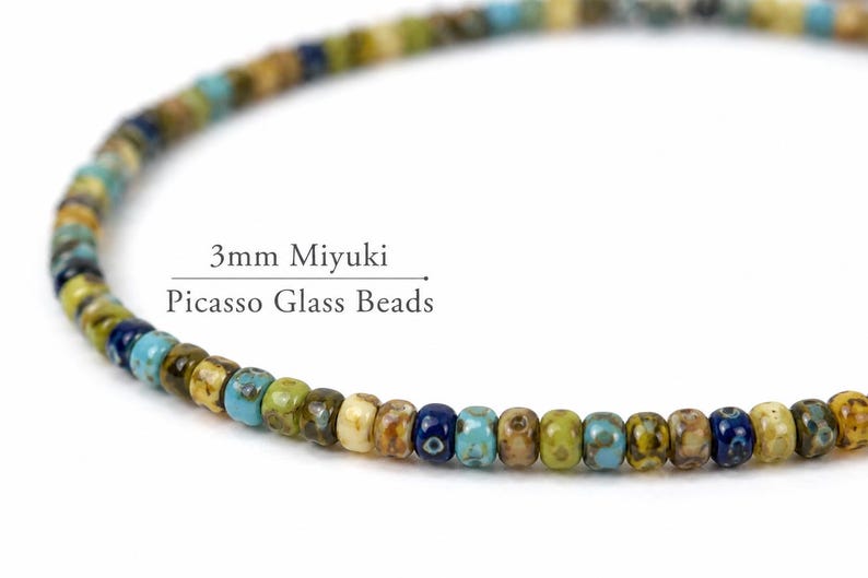 May include: A close-up of a strand of 3mm Miyuki Picasso glass beads. The beads are a mix of colors, including blue, green, yellow, and brown. The text "3mm Miyuki Picasso Glass Beads" is visible.