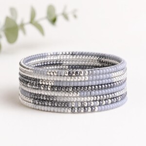 Könnte beinhalten: Ein mehrreihiges Armband aus mehreren Reihen kleiner, runder Perlen in den Farben Hellgrau, Dunkelgrau und Metallic-Silber. Die Perlen sind in einem sich wiederholenden Muster angeordnet und bilden eine gestapelte, kreisförmige Form.